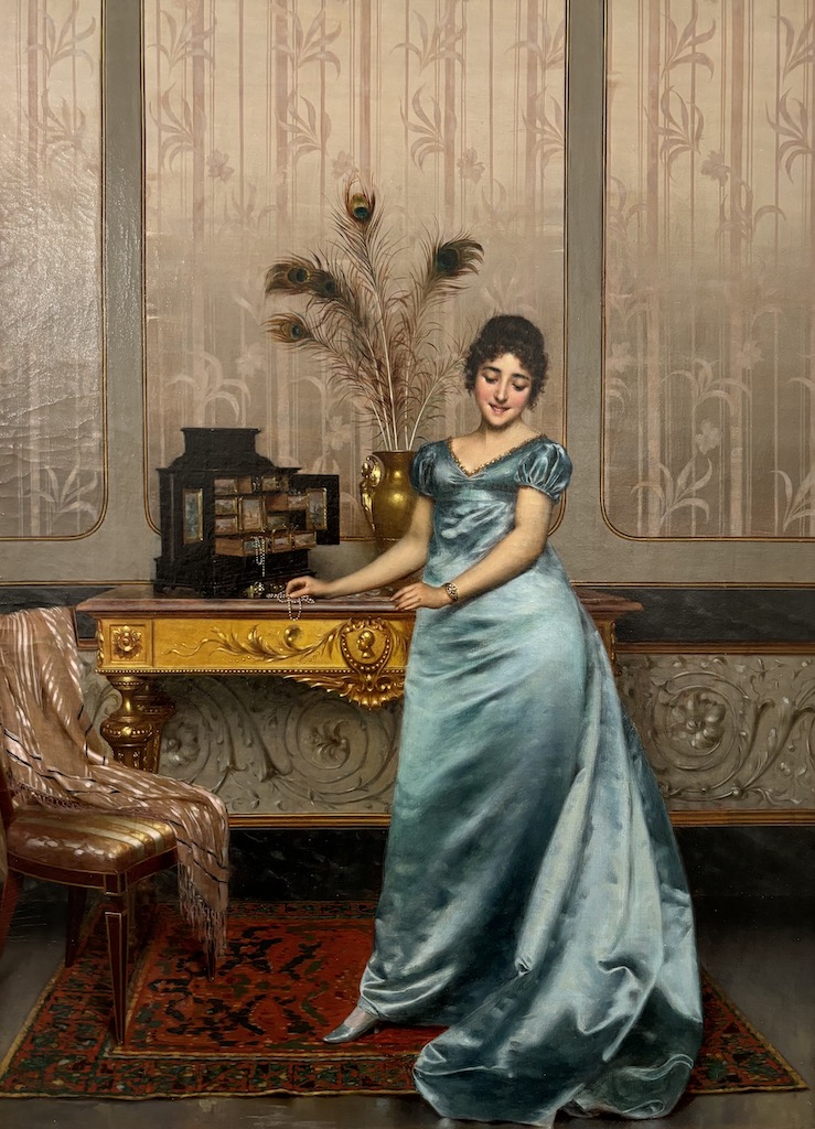 Bashful Model by Vittorio Reggianini depicts a pale women wearing a cyan blue dress that's drapping on the carpet below. The women stands infront of a gold desk and open jewellery box.